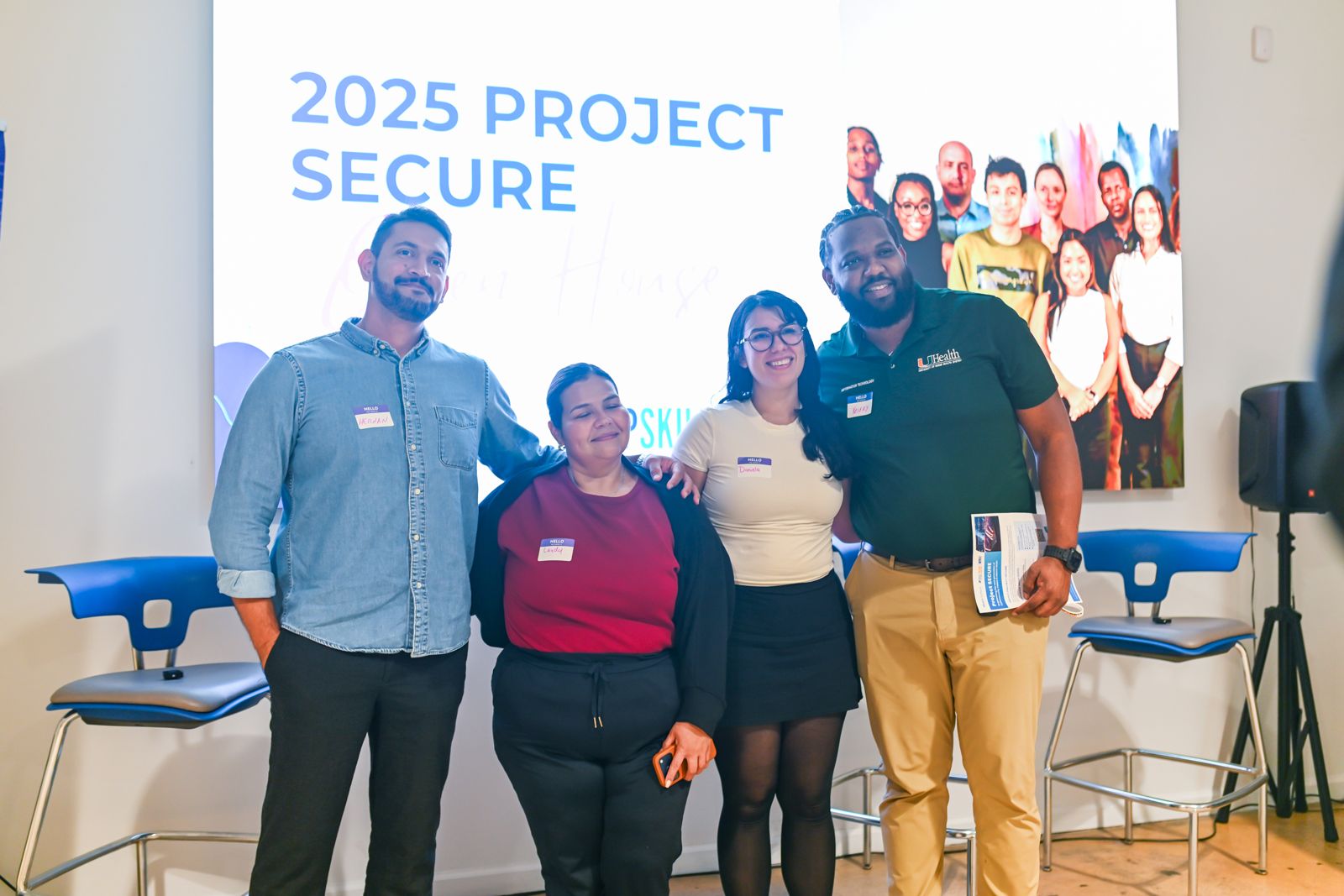 Four Project SECURE participants smiling in front of a 2025 Project SECURE open house slide